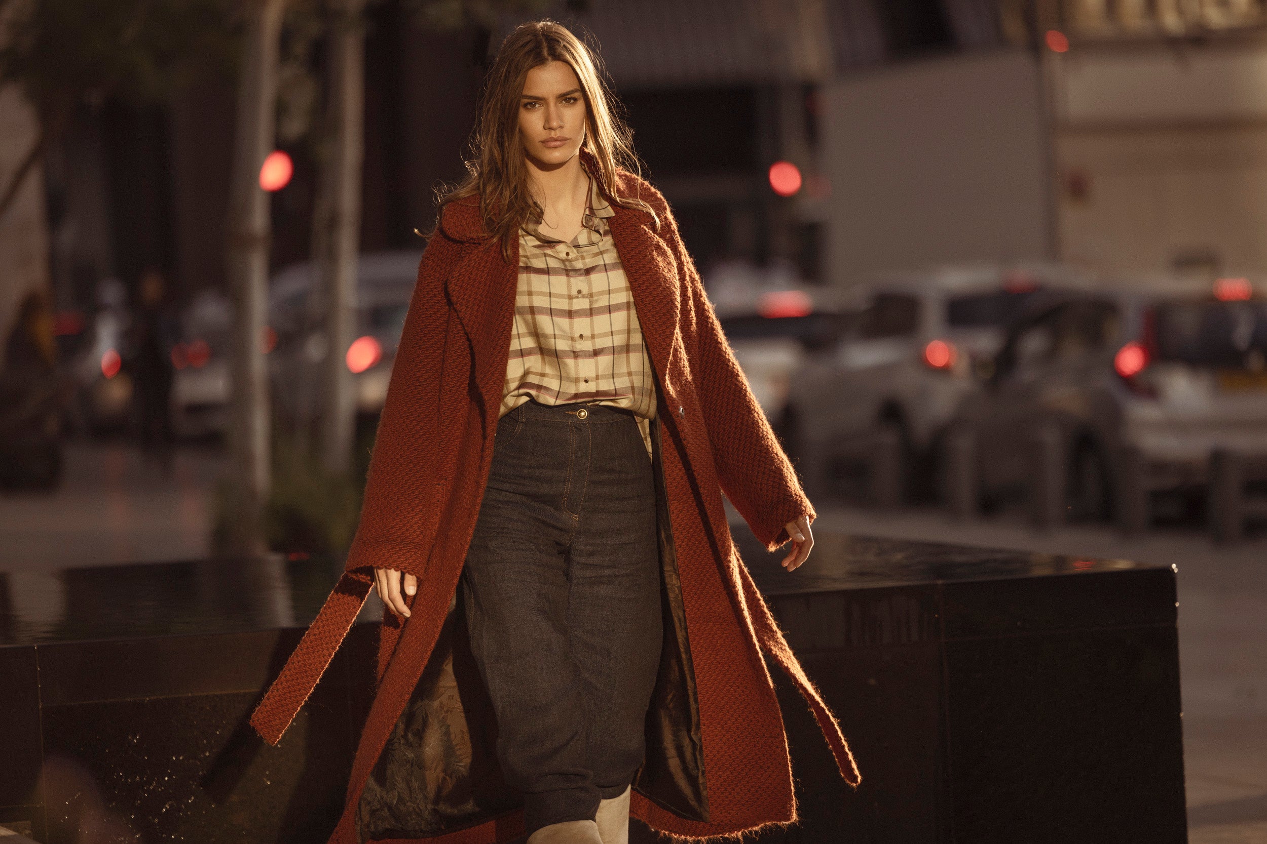 A model wears long woolen coat in color brick with belt and front pockets  